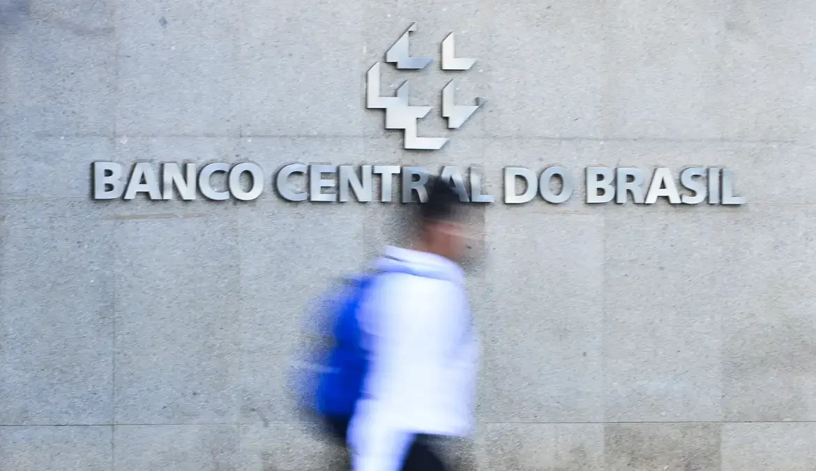 A foto mostra a fachada do Banco Central do Brasil. Um homem passa em frente