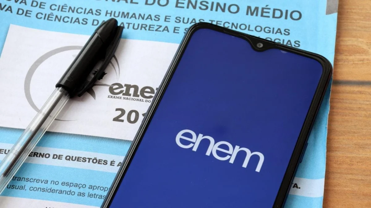 A foto mostra um caderno de provas do Enem na cor azul, uma caneta esferográfica na cor preta e um celular com a tela azul marinho escrito