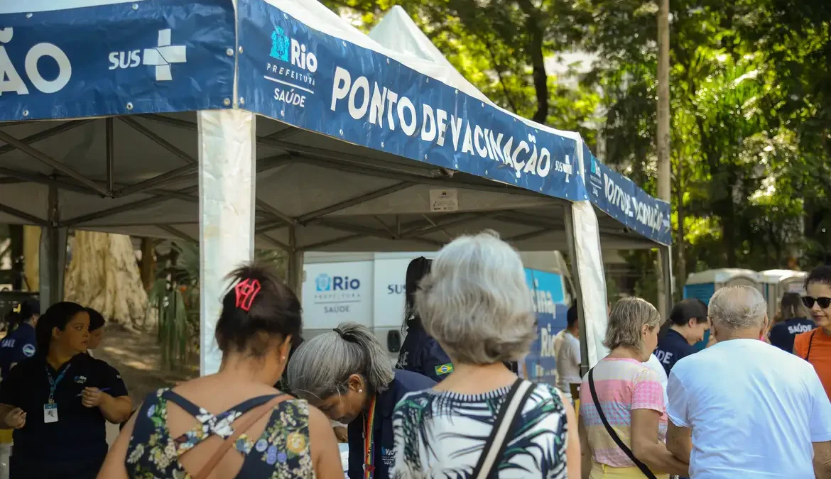 A foto mostra uma tenda que funciona com ponto de vacinação. Tem várias pessoas aguardando para se vacinar. A foto foi tirada de dia